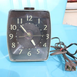 May include: A vintage brown alarm clock with a black face and white numbers. The clock has a cord with a plug.