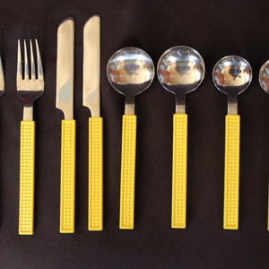 May include: A set of eight stainless steel flatware pieces arranged in a row, consisting of two forks, two knives, two large spoons, and two small spoons. Each piece features a bright yellow handle with a textured, grid-like pattern.