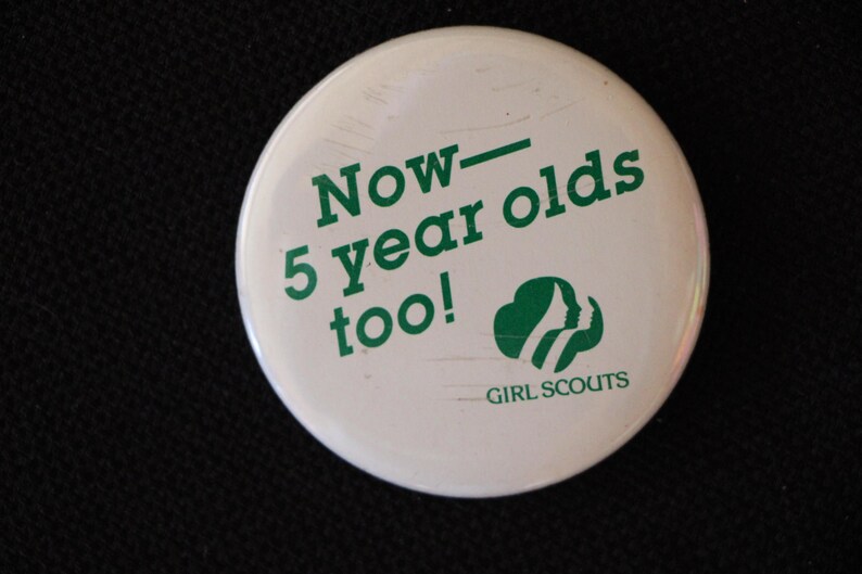 May include: White button with green text that reads "Now - 5 year olds too!" and a green Girl Scout logo.