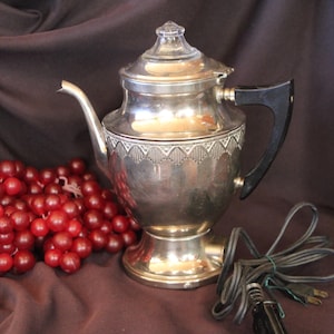 May include: A silver-toned electric percolator with a black handle and a clear glass knob on the lid. The body features a decorative etched pattern around the upper section. A black power cord is attached to the base of the metallic vessel.