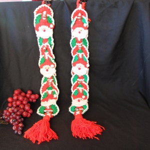May include: Two crocheted Christmas garlands with red, white, and green Santa Claus figures and bells. Each garland has a red tassel at the bottom.