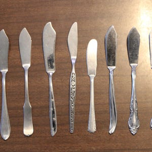 May include: An assortment of eight silver butter knives, each with a unique blade shape and handle design. The knives are arranged on a dark brown wooden surface, showcasing a variety of vintage and modern styles.