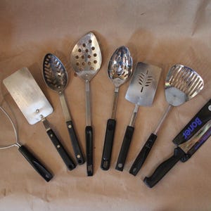 May include: A set of seven kitchen utensils, including a potato masher, a spatula, a slotted spoon, a ladle, a slotted turner, a solid spoon, and a knife. The utensils have black handles and are made of stainless steel.