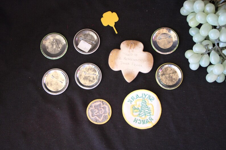 May include: A collection of Girl Scout badges and pins, including a round badge with a green four-leaf clover and the text "Girl Scout" and a round badge with a green tree and the text "Zynk Park Branch".