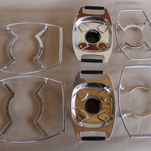 May include: Four silver-toned metal tea pot stands. Two of the stands are round with a center hole and a black handle. The other two stands are oval shaped with a curved design.