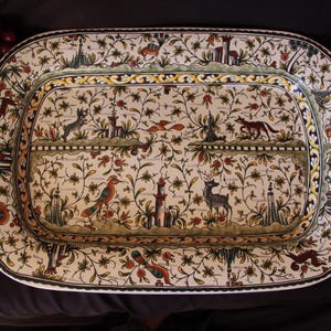 May include: A large rectangular ceramic serving platter with rounded corners, featuring a cream-colored background decorated with intricate hand-painted illustrations of various animals, birds, towers, and dense floral vines in shades of green, red, and brown.