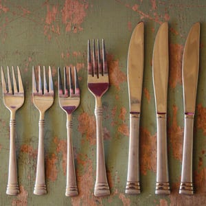 May include: A collection of vintage silverware, including forks and knives, arranged on a distressed green surface. The forks have four tines, and the knives have a simple, elegant design. The silverware is made of a shiny, metallic material.