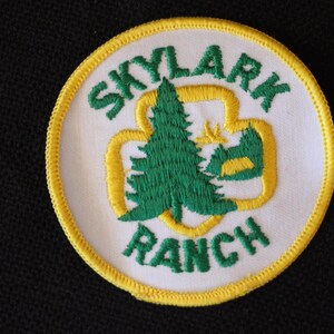 May include: A white embroidered patch with a yellow border. The patch features a green pine tree inside a yellow four-leaf clover design. The text "SKYLARK RANCH" is embroidered in green around the design.