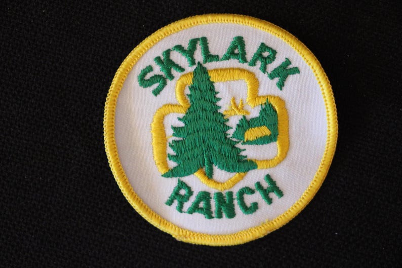 May include: A white embroidered patch with a yellow border. The patch features a green pine tree inside a yellow four-leaf clover design. The text "SKYLARK RANCH" is embroidered in green around the design.