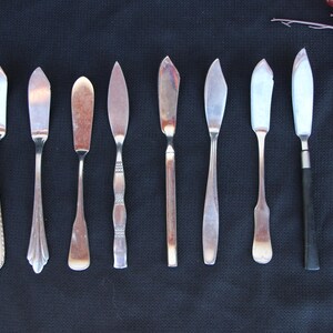 May include: An assortment of butter knives with various handle and blade designs. The knives are made of silver-colored metal and are arranged on a dark background. Some handles have decorative patterns, while others are plain.