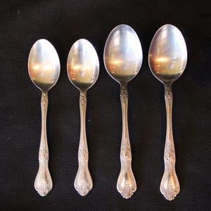 May include: Four silver spoons of varying sizes are arranged vertically on a black surface. The spoons have ornate, detailed handles and oval bowls. The spoons are likely for serving or eating.