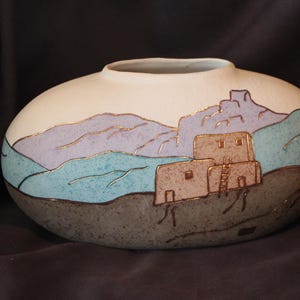 May include: A low, oval-shaped ceramic vase with a matte finish. The surface features a painted landscape with a tan adobe-style building, purple mountains, and a teal foreground, all outlined in metallic gold paint against a cream background.
