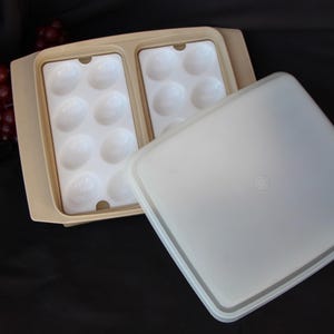 May include: A beige egg container with a clear lid. The container has two sections, each holding six white egg-shaped molds. The lid is off to the side. A bunch of red grapes is in the background.