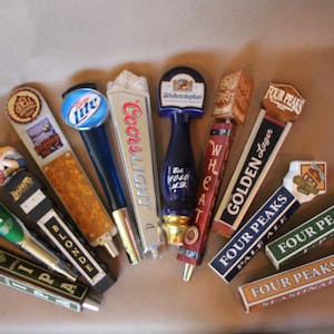 May include: A collection of colorful beer tap handles with various brand names, including Coors Light, Heineken, and Four Peaks. The handles are made of metal and plastic and feature different designs.