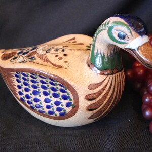 May include: A ceramic duck figurine with a detailed, hand-painted design. The duck features a cream-colored body with blue, brown, and green accents. The duck's head is painted with blue, green, and brown, and it has a white band around its beak. The duck is a decorative item.