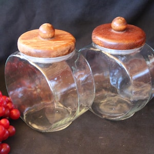 May include: Two clear glass jars with wooden lids. The jars have a unique, curved shape and are topped with round, wooden knobs. The lids are a warm brown color. A cluster of red berries is visible on the left side of the image.
