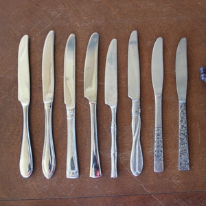 May include: Eight silver dinner knives with different handle designs, laid out on a wooden surface. A bunch of dark purple grapes is on the right side of the image.