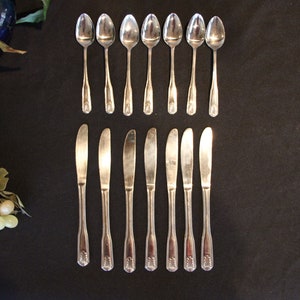May include: A set of 12 silver-toned flatware pieces, including 6 spoons and 6 butter knives, arranged in two rows on a black surface.