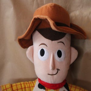 Puede incluir: Un juguete de peluche de Woody de la franquicia Toy Story. Lleva un sombrero de vaquero marr&oacute;n y una camisa de cuadros amarillos y rojos. Tiene una expresi&oacute;n amigable en su rostro.