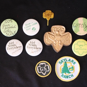 May include: A collection of Girl Scout badges and pins, including a gold-colored Girl Scout pin, a green and yellow badge that says "Yes on Girl Scouting", a green and white badge that says "Now - 5 year olds too!" and a green and white badge that says "100% Green blooded GIRL SCOUT".