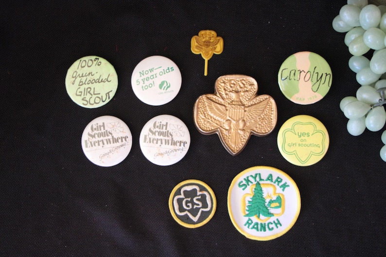 May include: A collection of Girl Scout badges and pins, including a gold-colored Girl Scout pin, a green and yellow badge that says "Yes on Girl Scouting", a green and white badge that says "Now - 5 year olds too!" and a green and white badge that says "100% Green blooded GIRL SCOUT".