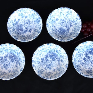 May include: Five white and blue floral patterned bowls. The bowls have a scalloped edge and a delicate blue floral design. The bowls are arranged on a dark surface, with a cluster of red berries in the background.