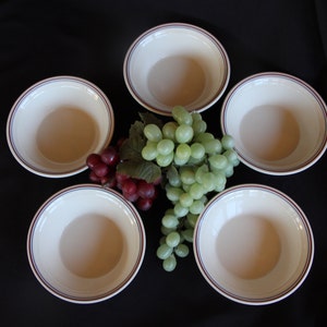 May include: Five white bowls with a red and blue stripe around the rim. The bowls are arranged in a circle with a bunch of green and red grapes in the center.