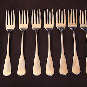 May include: A set of seven identical silver-toned metal forks arranged in a parallel row. Each fork features a long, slender handle with a slightly flared, rounded end and four tines. The polished metallic surface reflects light evenly across the set.