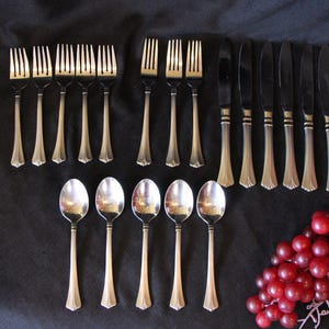 May include: A collection of silverware, including forks, knives, and spoons, arranged on a dark surface. The silverware has a silver finish with decorative handles. A bunch of red grapes is in the lower right corner.