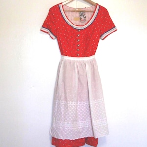 May include: A red and white floral print dress with a white apron. The dress has a scoop neckline with blue trim and short sleeves. The apron is white with a lace hem. The dress is hanging on a wooden hanger.