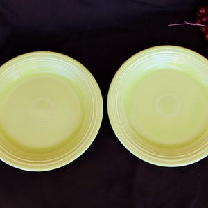 May include: Two pale yellow ceramic plates with concentric ring detailing. The plates are round with a slightly raised rim and a central circular indentation. The background is a dark, solid color, with a cluster of red grapes in the upper right corner.