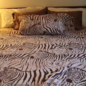 Puede incluir: Una colcha y una funda de almohada a juego con un patr&oacute;n repetitivo de rayas de cebra marrones y blancas intercaladas con sutiles ilustraciones de caras de tigre. La ropa de cama est&aacute; dispuesta sobre una cama con almohadas lisas.