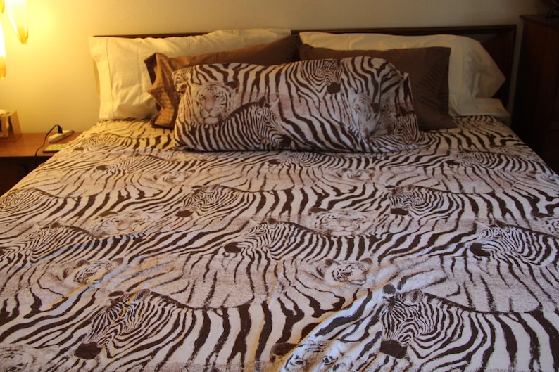 Puede incluir: Una colcha y una funda de almohada a juego con un patr&oacute;n repetitivo de rayas de cebra marrones y blancas intercaladas con sutiles ilustraciones de caras de tigre. La ropa de cama est&aacute; dispuesta sobre una cama con almohadas lisas.