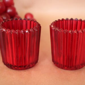 May include: Two cylindrical votive candle holders made of translucent red glass. Each holder features a vertical ribbed texture along the exterior walls and a scalloped, serrated top rim. The glass is deep red, allowing light to pass through.
