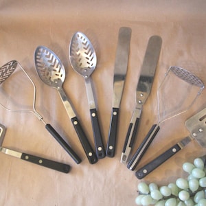 May include: A collection of vintage kitchen utensils, including a slotted spoon, spatula, and masher, with black handles and stainless steel heads. The tools are arranged on a light brown surface.