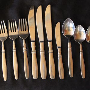May include: A collection of silverware including forks, knives, and spoons. The utensils have light-colored handles with a dark band near the top and bottom. The blades and tines are silver-colored.