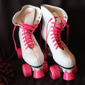 May include: A pair of white quad roller skates featuring bright pink laces and matching pink wheels. The skates have a high-top boot design with metal eyelets for the laces. The brand name Penfield is printed in pink on the tongue of the boot.