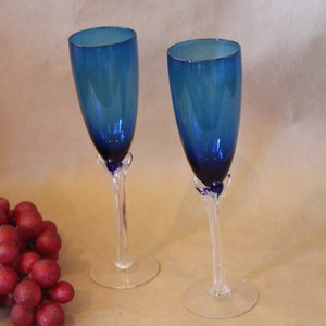 May include: Two blue glass champagne flutes with clear stems. The flutes have a gradient of blue from dark to light.