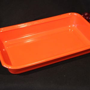 May include: Bright orange rectangular tray with rounded corners. The tray is empty and has a glossy finish. A bunch of red grapes is visible in the background. The tray is likely used for serving or storage.