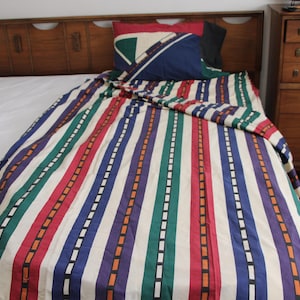 May include: A bed with a wooden headboard and a colorful striped comforter. The comforter features vertical stripes in red, green, blue, purple, and orange, separated by white lines. Pillows with geometric designs are on the bed. A wooden dresser is in the background.