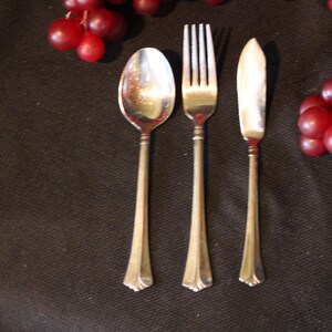 May include: Silverware set including a spoon, fork, and knife, arranged on a dark surface. The utensils have long, slender handles and decorative ends. Red grapes are in the background.
