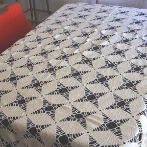 May include: Cream-colored, openwork tablecloth with a repeating starburst pattern. The tablecloth is draped over a rectangular table, with the pattern visible across the surface. The design features a series of interconnected star shapes.