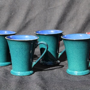 May include: Four teal-colored ceramic mugs with a blue interior and a white rim. Each mug has a handle and a flared shape. The mugs are arranged on a dark surface, with a cluster of red grapes in the background.