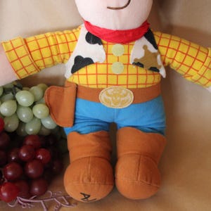 Puede incluir: Un juguete de peluche de Woody de la franquicia Toy Story. El juguete lleva una camisa de cuadros amarillos y rojos, un chaleco marr&oacute;n, un cintur&oacute;n azul y pantalones vaqueros azules. El juguete tiene una bandana roja alrededor del cuello y una estrella de sheriff en el pecho.