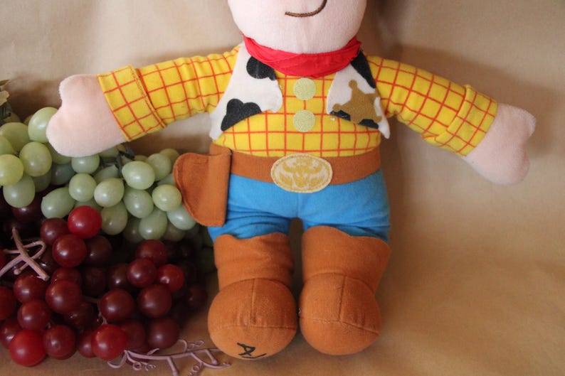 Puede incluir: Un juguete de peluche de Woody de la franquicia Toy Story. El juguete lleva una camisa de cuadros amarillos y rojos, un chaleco marr&oacute;n, un cintur&oacute;n azul y pantalones vaqueros azules. El juguete tiene una bandana roja alrededor del cuello y una estrella de sheriff en el pecho.