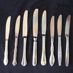 May include: A set of eight silver butter knives with different handle designs. The knives are arranged in a row on a black surface.