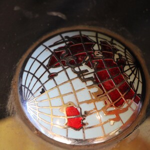 Puede incluir: Un emblema redondo con un dise&ntilde;o de mapa del mundo. El globo terr&aacute;queo est&aacute; representado en rojo y dorado sobre un fondo blanco, con un patr&oacute;n de cuadr&iacute;cula. El emblema est&aacute; enmarcado en un c&iacute;rculo.