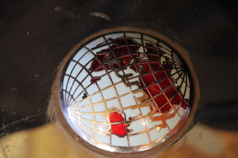 Puede incluir: Un emblema redondo con un dise&ntilde;o de mapa del mundo. El globo terr&aacute;queo est&aacute; representado en rojo y dorado sobre un fondo blanco, con un patr&oacute;n de cuadr&iacute;cula. El emblema est&aacute; enmarcado en un c&iacute;rculo.