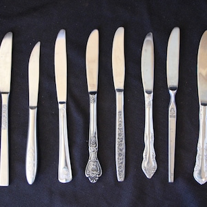 May include: Eight silver butter knives with different handle designs. The knives are arranged in a row on a black surface.