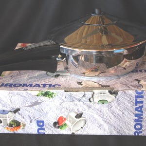 May include: A stainless steel pressure cooker with a black handle and a clear lid, resting on a box with the text "DUROMATIC" and "KUHN RIKON SWITZERLAND". The cooker is reflective, and the box has illustrations of the product.